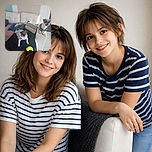 Two adorable small dogs wearing cozy sweaters are chilling on a white couch and its armrest, surrounded by pillows and a plush toy, with a bright green toy in the foreground. The photo has a casual homey vibe but is slightly blurry.