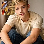 A tiny chihuahua puppy peeks up from a plaid blanket with comically large ears and soulful eyes, sharing a cozy couch spot beside a relaxed person — looks like it's plotting how to get extra treats.