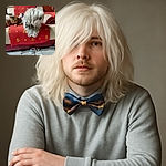A fluffy white dog with a checkered bandana sits on a red-patterned couch cover, next to a relaxed tabby cat also lounging comfortably. A single sandal lies on the tiled floor in front, adding a casual homey vibe to the cozy scene.