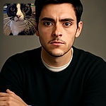 A close-up photo of a curious black and white cat with large eyes and prominent whiskers, looking directly into the camera with a slightly blurred background that hints at a cozy indoor setting.