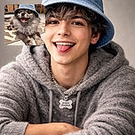 A fluffy dog sporting a cool denim bucket hat and a chain necklace with a bone-shaped tag that reads 'papa maman' lounges happily on a patterned rug, tongue out and clearly enjoying the spotlight indoors.