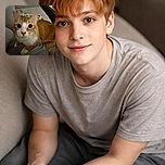 A curious orange and white cat with big round eyes stares directly into the camera, perched on a cozy couch with a person lounging in the background. The intimate close-up captures the cat's whiskers and pink nose in soft lighting, creating a warm and homely atmosphere.