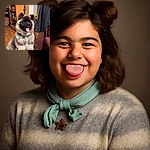 A fluffy dog with a thick coat and a mint-green scarf around its neck sits happily indoors, tongue out and looking content. The cozy room features wooden flooring, a bookshelf filled with various items, a sunflower bouquet in the background, and a jacket hanging on a chair.