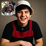 A happy black and white dog wearing a white hat and red harness is lying on sandy ground, smiling with its mouth open. A person's hand is gently touching the dog's head, and a stroller and some shoes are visible in the background, giving a cozy beach day vibe.