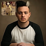 A serious-faced Boston Terrier sits attentively on a fluffy gray rug, framed by a cozy room with a wooden table leg, a yellow plastic bin, and a clear plastic cup filled with water nearby.