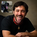 A happy black dog with a red collar is lying on a textured carpet, tongue hanging out in a joyful pant, with a cozy indoor setting featuring a door and some furniture in the background.