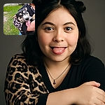 A happy puppy with one blue eye and one brown eye is being held by a person wearing a black and leopard print top, tongue out and looking playful, with a grassy background and a dirt path.