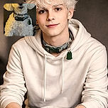 A fluffy white dog with a green collar sits attentively against a dark background, looking curious and ready for a treat. The lighting highlights the soft curls of its fur, while a small text overlay 'Crop' appears in the top corner, adding a quirky touch to the otherwise cozy portrait.