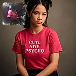 A charming black dog sits on a cozy couch, wearing a bright pink shirt that cheekily reads 'CUTE BUT PSYCHO'. The dog looks calm yet curious, with soft natural light filtering in from a window behind, casting gentle shadows and adding warmth to the scene.