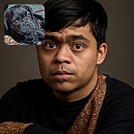 A close-up of a black pug puppy with big, shiny eyes looking curiously off-camera, lying comfortably on a patterned blanket and soft cushions, with a cozy indoor vibe.