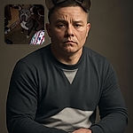 A serious-looking dog with glowing eyes from the flash sits on a worn carpet inside a dimly lit room cluttered with household items, including a laundry basket and shoes, while a colorful patterned blanket peeks into the frame.