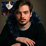 A charming black and white cat wrapped in shiny blue and black tinsel poses regally on a red and gray fabric surface with a tall orange vase in the background, looking curiously at the camera with bright green eyes.