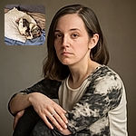 A charming dachshund with a unique merle pattern is cozily curled up on a white pillow atop a blue blanket on a gray couch, looking attentively at the camera with one ear flopped over.
