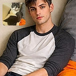A chubby gray and white cat lounges comfortably against a wall, looking directly at the camera with a calm and slightly curious expression, surrounded by cozy blankets and household items.