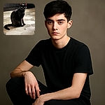 A sleek black cat sits calmly on a patterned hexagonal tile floor, its bright eyes staring directly at the camera with a curious yet mysterious gaze. The dark cabinetry in the background contrasts with the lighter floor, highlighting the cat's shiny fur.