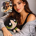 A charming dog lounges on a cozy blanket, hugging a plush toy with big green eyes, looking off-camera with a thoughtful expression, surrounded by a casual bedroom setting.