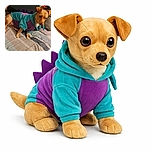 A small dog dressed in a colorful dinosaur costume stands on a bed with a patterned blanket. The dog's expression is curious and slightly cautious, while the background is softly blurred, focusing attention on the adorable costume and the dog's stance.