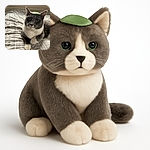 A charming gray and white cat lounges comfortably on a patterned bedspread with a small green leaf playfully balanced on its head, looking curiously at the camera with bright green eyes. The background shows a windowsill with various potted plants, adding a cozy indoor vibe.