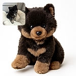 A fluffy little dark-furred puppy with soulful eyes lounges comfortably on a soft, white, plush blanket, looking curiously at the camera with a cozy and heartwarming backdrop.