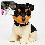 A charming dog with a black, brown, and white coat lounges comfortably on a couch, resting against a pink pillow with a floral design. The dog wears a black and white patterned collar and gazes attentively at the camera with soulful eyes, while the background features a gray couch with visible stitching.