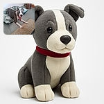 A playful dog with a gray and white coat is lying on a patterned cushion, mouth wide open as if mid-yawn or mid-bark, with a cozy beige rug and an African drum in the background adding a homely vibe.