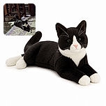 A tuxedo cat lounges dramatically on a patterned rug beneath a forest of wooden chair legs, looking like it has claimed the dining room throne and is waiting for its royal snack delivery.