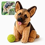 A playful German Shepherd dog lies on the grass with its head tilted adorably, tongue out, and a well-used tennis ball between its paws, surrounded by lush greenery and a tree line in the background.