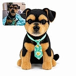 A serious little dog sits proudly on a turquoise blanket, wearing a colorful flower tag and giving the camera a stare that says, “I supervise Christmas now.” A decorated tree, couch, framed picture, and cozy living room complete the scene.