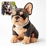 A sturdy French Bulldog basks in the sunlight on a stone surface with a backdrop of wildflowers and greenery, looking curiously at the camera with its large ears perked up.