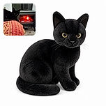 A sleek black cat lounges like a tiny fireplace inspector, staring intensely beside a glowing wood stove while wrapped in a richly patterned red-and-gold blanket. The cozy scene says maximum warmth, minimum responsibility.