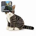 A striped cat sits like the neighborhood’s tiny security guard, staring intensely down a fenced walkway while its dramatically long tail stretches across the scene. Bright blue sky, sunny buildings, artificial grass, and scattered leaves complete the outdoor courtyard setting.