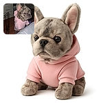 A small dog with a brindle coat and large ears is standing on its hind legs, wearing a cozy pink hoodie, looking curiously over a gray surface in a dimly lit room with wooden floors and some furniture.