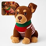 A charming dog wearing a festive red, white, and green striped sweater sits patiently on a couch adorned with a colorful quilt. Behind, a decorated Christmas tree with glowing lights and red ornaments adds a warm holiday ambiance, while a stocking hangs on a chair against a rustic brick wall.