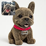 A close-up photo of a happy French Bulldog with a pink collar sitting inside a car, looking excited and ready for an adventure. The background is softly blurred, focusing all attention on the dog's expressive face and big ears.