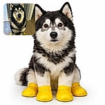 A fluffy dog with a striking black and white coat is sitting indoors wearing bright yellow booties on all four paws, looking up with a curious and slightly confused expression on its face. The background shows a kitchen and hallway with wooden flooring.