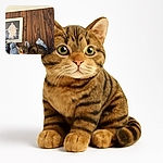 A curious tabby cat with wide eyes is perched among blue and white ceramic vases and a cat-shaped figurine, set against a rustic wooden wall with a framed picture of a statue in the background.