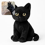 A sleek black cat with bright yellow eyes sits curled up on a colorful patterned rug, looking intently at the camera with a curious and slightly mysterious expression. The wooden floor and white cabinets in the background add a cozy indoor atmosphere.