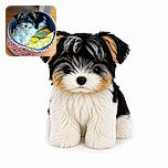 A fluffy black, white, and tan puppy lounges proudly in a colorful cushioned bed, looking like a tiny royal who has claimed the comfiest throne in the house. The bright yellow blanket and patterned sides add cheerful charm against the warm wooden floor.