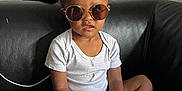 Isaac a rejoint le concours — aidez-le/la à gagner de superbes lots ! toddler, child, sunglasses, white_onesie, black_couch, keys, remote_control, curly_hair, bracelet, necklace, indoor, wallpaper, patterned_wall, sitting, cross_legged, brown_skin, cute, casual, home, portrait