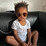 toddler, child, sunglasses, white_onesie, black_couch, keys, remote_control, curly_hair, bracelet, necklace, indoor, wallpaper, patterned_wall, sitting, cross_legged, brown_skin, cute, casual, home, portrait