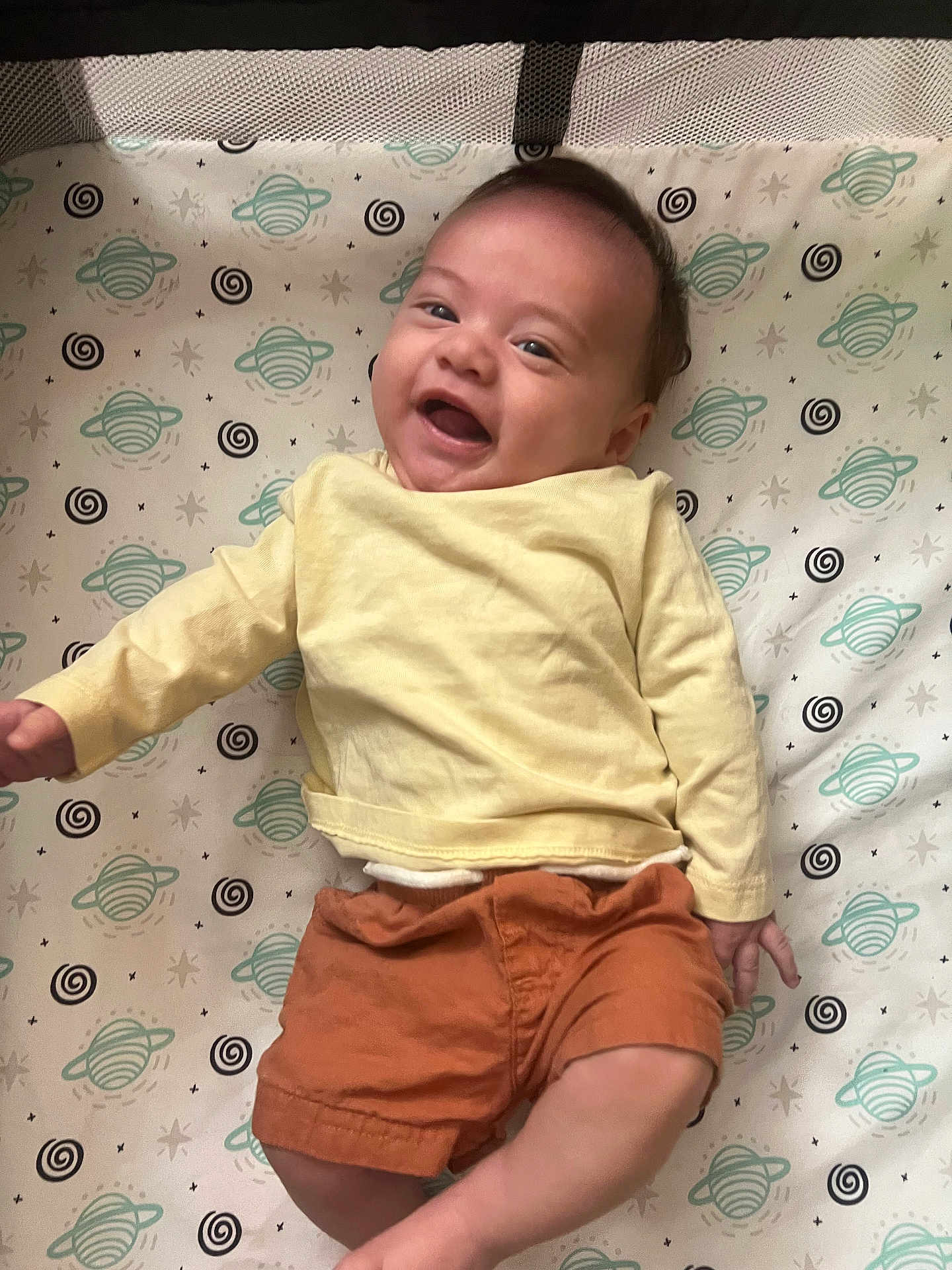 Jackson is registered to the contest to win money with this photo: baby, smiling, infant, yellow_shirt, orange_shorts, lying_down, blanket, pattern, space_theme, planets, stars, happy, child, cute, portrait, indoors, skin, head, face, person
