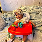 Valentina is registered to the contest to win money with this photo: baby, high_chair, toy, headband, bow, orange_tray, smiling, happy, indoor, blanket, pillow, footwear, white_clothing, child, person, seat, furniture, play, cute, infant