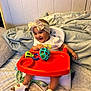 baby, high_chair, toy, headband, bow, orange_tray, smiling, happy, indoor, blanket, pillow, footwear, white_clothing, child, person, seat, furniture, play, cute, infant