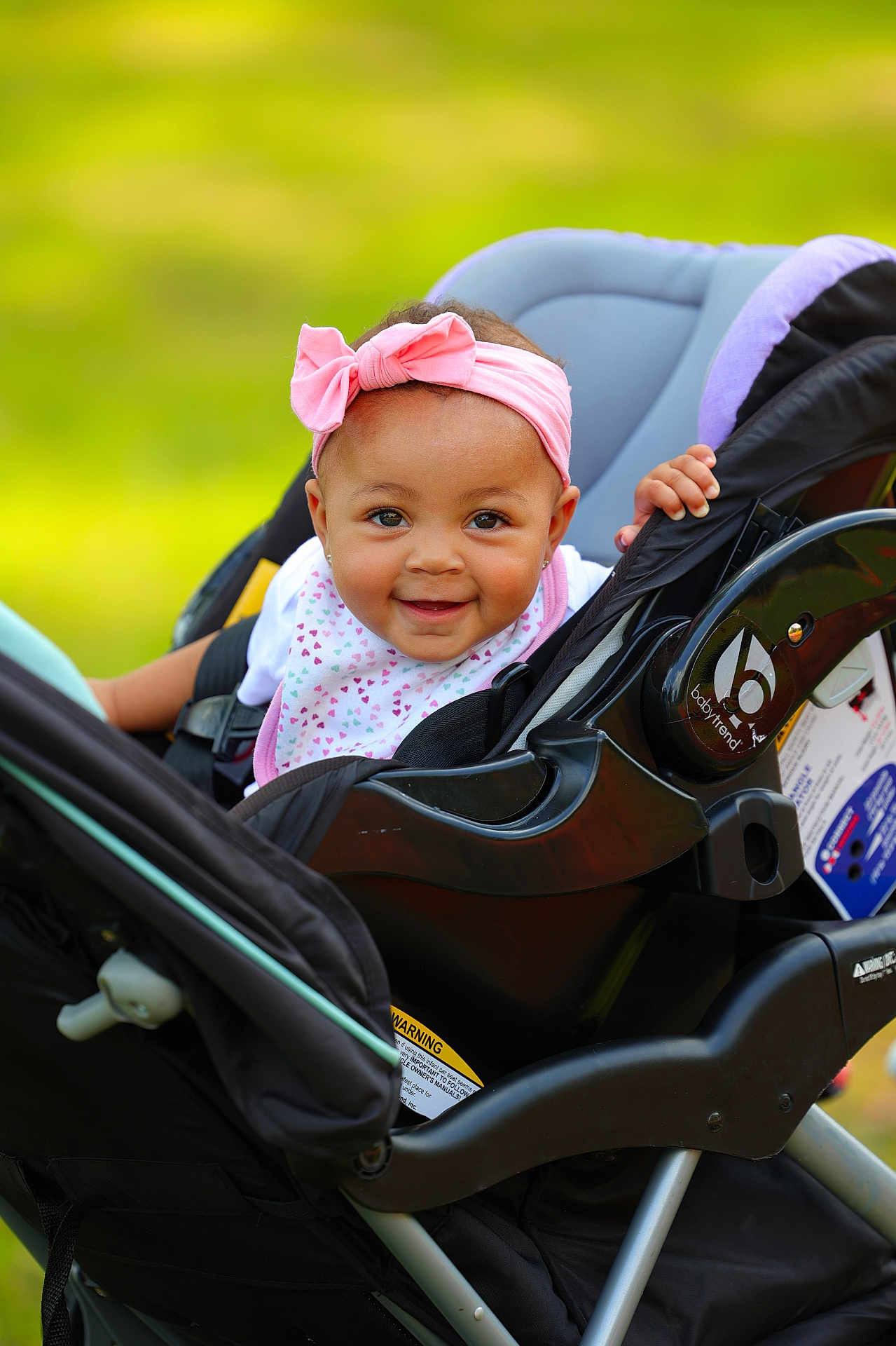 Brielle is registered to the contest to win money with this photo: baby, child, stroller, pink_bow, headband, smile, happy, outdoor, green_background, infant, clothing, seat, person, cute, young_child, baby_bib, hand, face, portrait, daylight