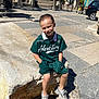 child, boy, smiling, outdoor, sunlight, stone_bench, pavement, green_clothing, sneakers, shorts, casual, urban, building, window_shutters, bench, daytime, shadow, happy, portrait, leisure