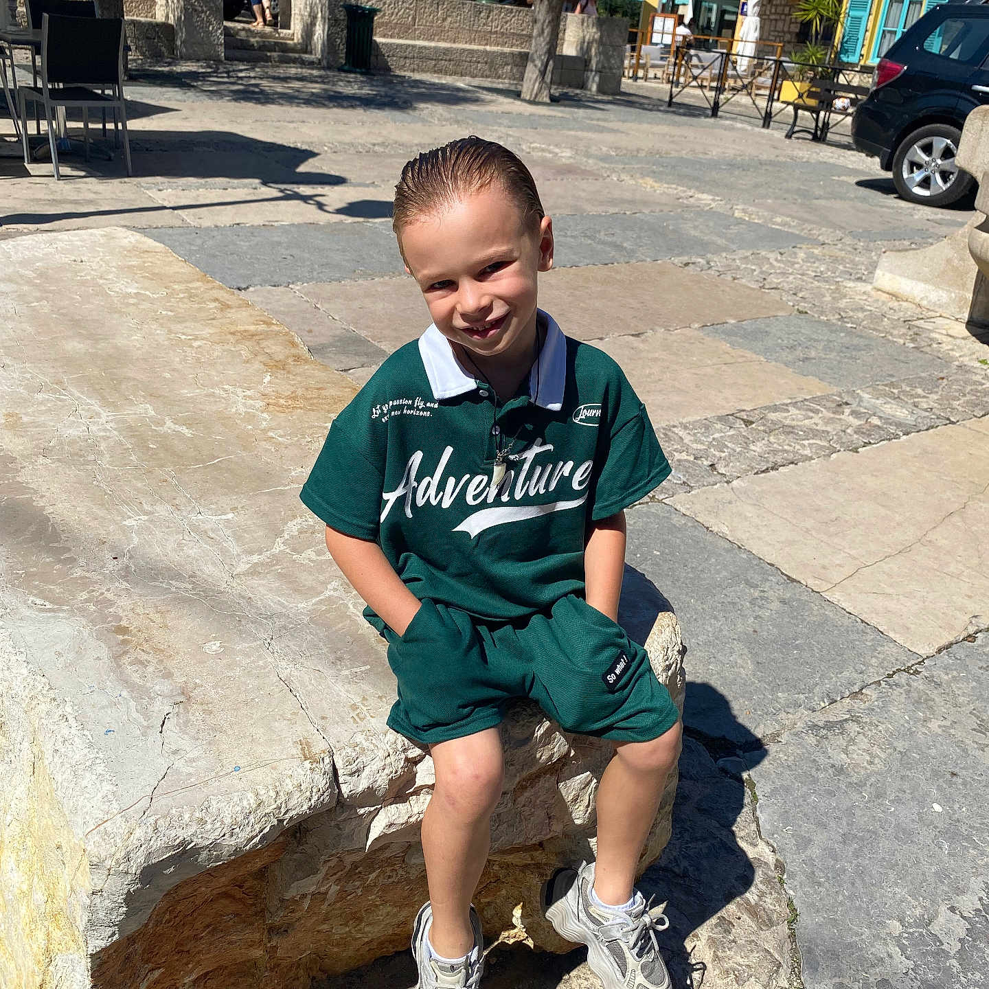 Enzio a rejoint le concours — aidez-le/la à gagner de superbes lots ! bench, boy, building, casual, child, daytime, green_clothing, happy, leisure, outdoor, pavement, portrait, shadow, shorts, smiling, sneakers, stone_bench, sunlight, urban, window_shutters