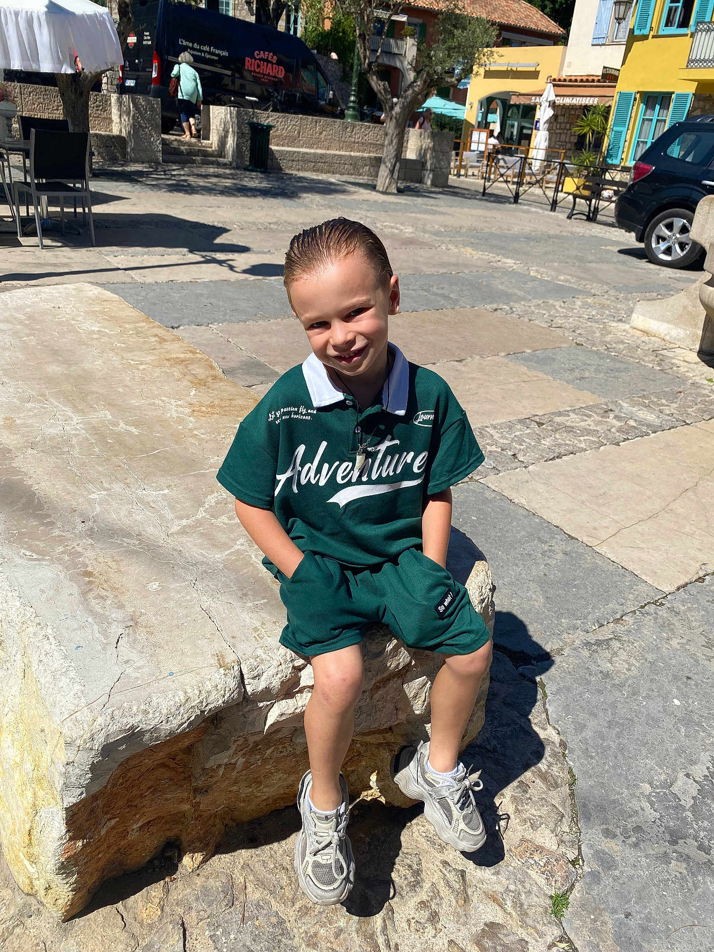 Enzio a rejoint le concours — aidez-le/la à gagner de superbes lots ! child, boy, smiling, outdoor, sunlight, stone_bench, pavement, green_clothing, sneakers, shorts, casual, urban, building, window_shutters, bench, daytime, shadow, happy, portrait, leisure