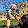 adult, baby, blue_sky, castle, child, clothing, fence, happy, holding, joy, outdoor, park, person, play, smile, stone_wall, sunlight, tattoo, tree, water