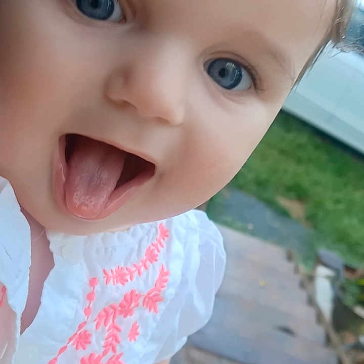 Wednesday is registered to the contest to win money with this photo: baby, blue_eyes, car, child, closeup, cute, daylight, embroidered_clothing, face, greenery, headshot, infant, outdoor, patio, person, playful, smiling, tongue_out, white_shirt, young_child