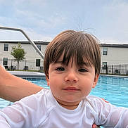 Isaac is registered to the contest to win money with this photo: toddler, child, swimming_pool, water, white_shirt, hand, building, sky, outdoor, residential, fence, support, portrait, cute, young_child, person, face, hair, expression, daytime
